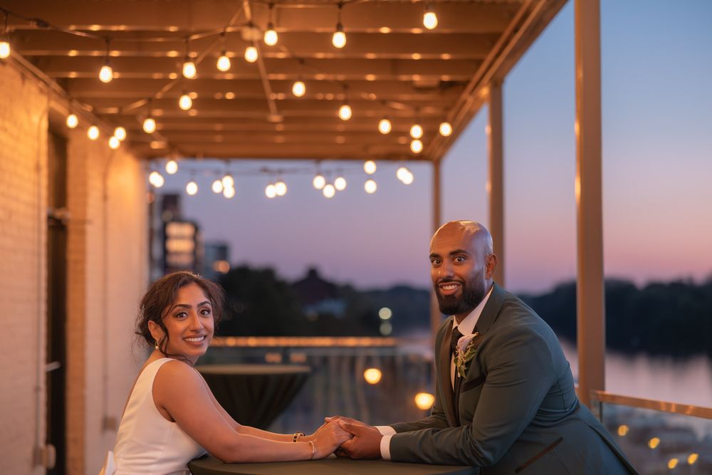 Bride and groom hold hands on a romantic rooftop at sunset, surrounded by string lights with a waterfront view.