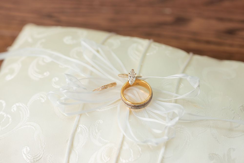 Gold wedding rings with diamond engagement ring resting on white satin ring bearer pillow with ribbon bow.