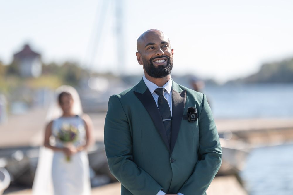 Smiling groom in green suit at waterfront wedding ceremony with bride in white dress in background.