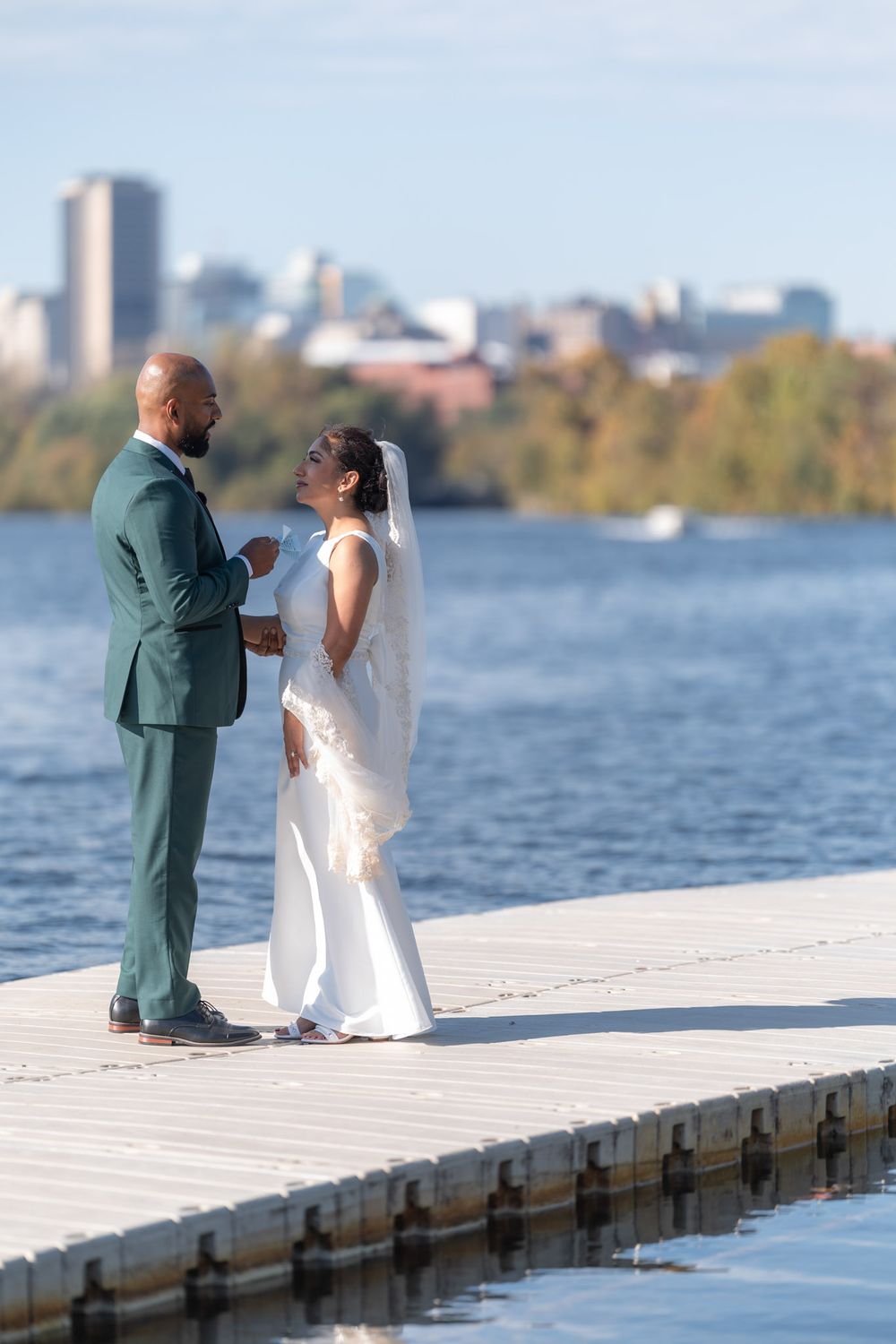 Bride and groom share a tender moment on a dock by the water with a city skyline backdrop on their wedding day.