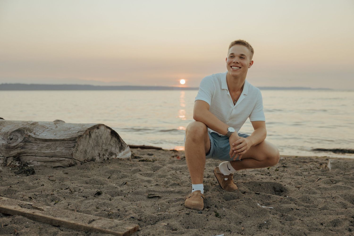 Young man smiling and crouching on sandy beach at sunset in Edmonds with driftwood and calm ocean waters in background.