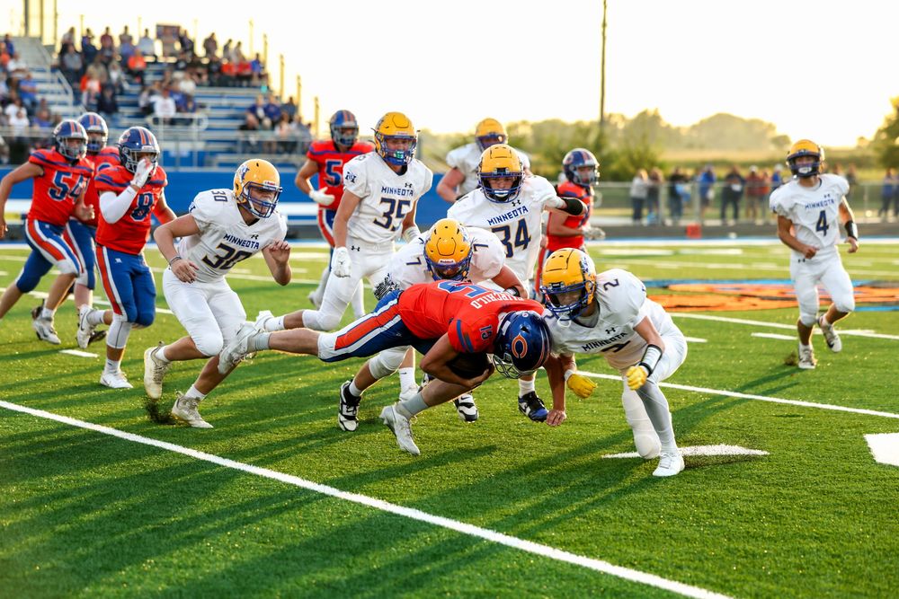High school football players in blue and gold uniforms tackle a ball carrier during an evening game on a green turf field.