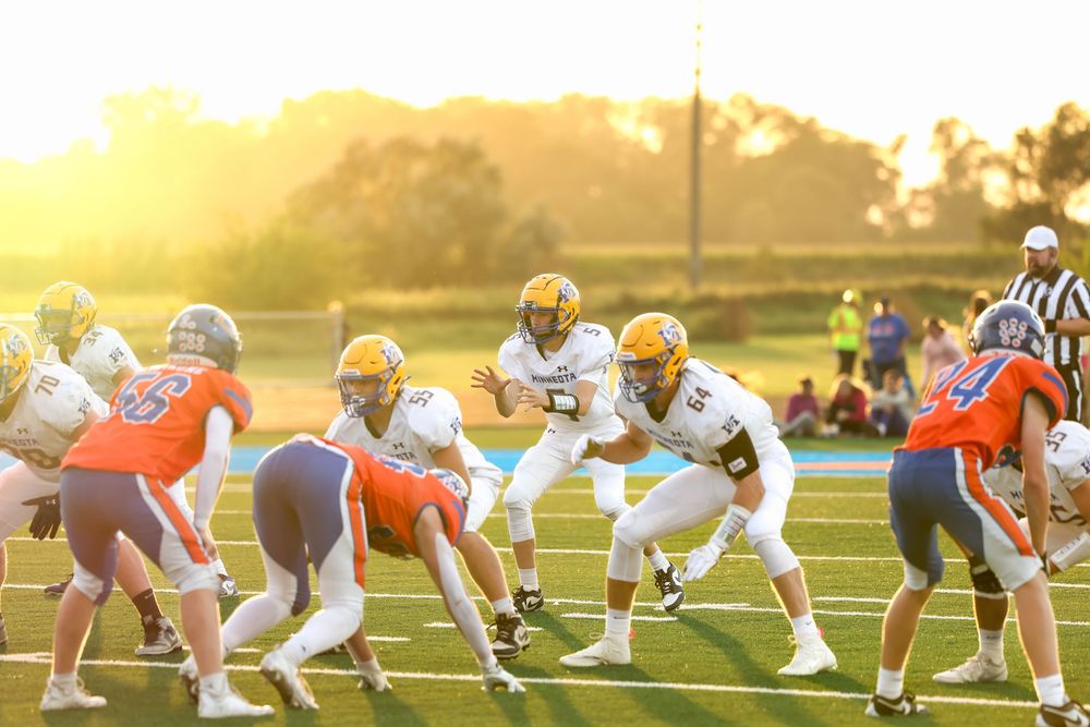 High school football players in gold uniforms line up against blue and orange opponents on a field at sunset.