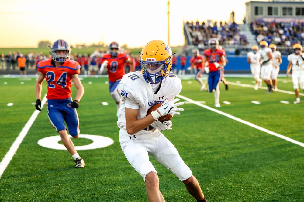 Football player in white and gold uniform catches ball during high school game on green turf field.