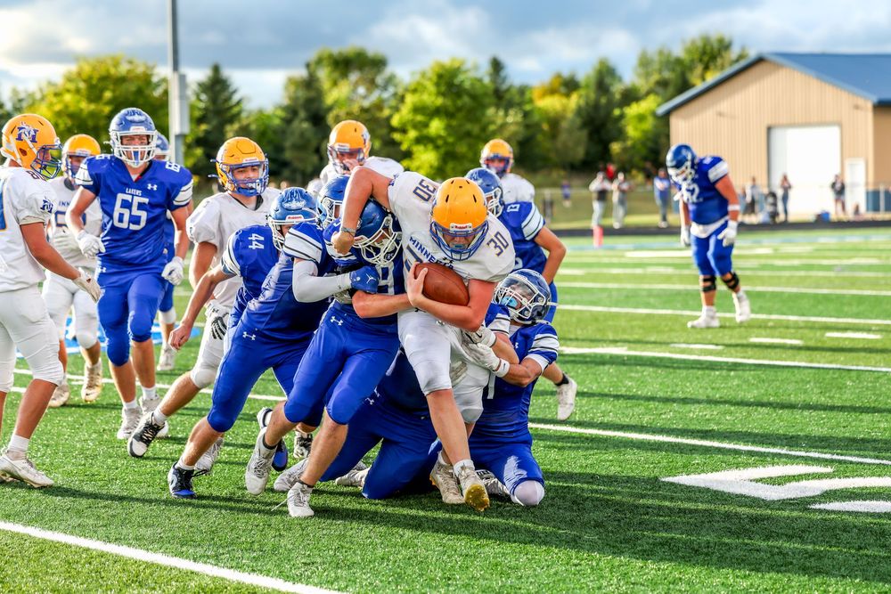 High school football players in blue and white uniforms tackle a ball carrier during an outdoor game on a green turf field.
