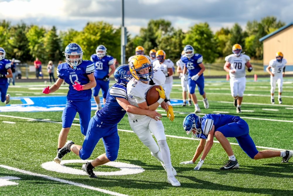 High school football player in white jersey runs with ball while blue-uniformed defenders attempt a tackle on green turf.