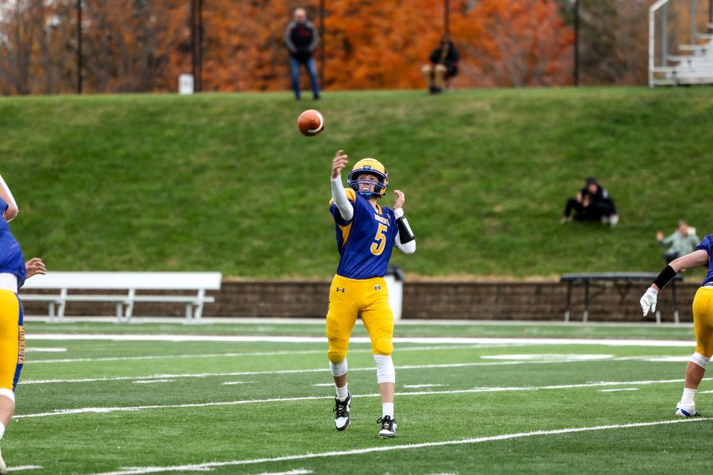 Football player in yellow and blue uniform throwing a pass during an outdoor game on a green turf field.