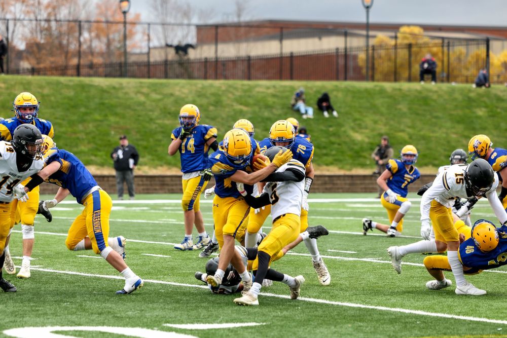 College football players in yellow and blue uniforms compete in an intense tackle during a daytime game on a green field.