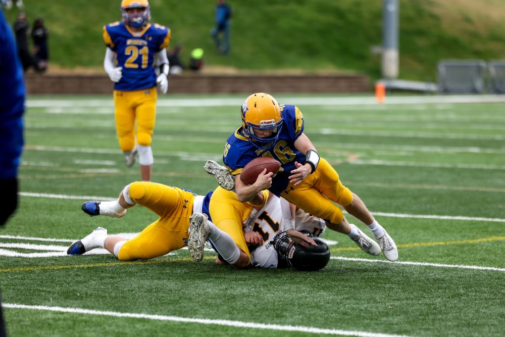 Football players in blue and gold uniforms tackle an opponent during an intense college football game on a green turf field.