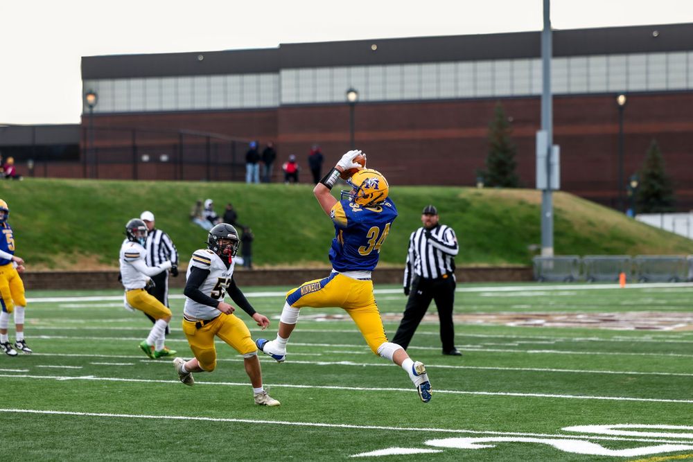 Football player in blue and gold jersey leaping to catch a pass during a college football game on a cloudy day.