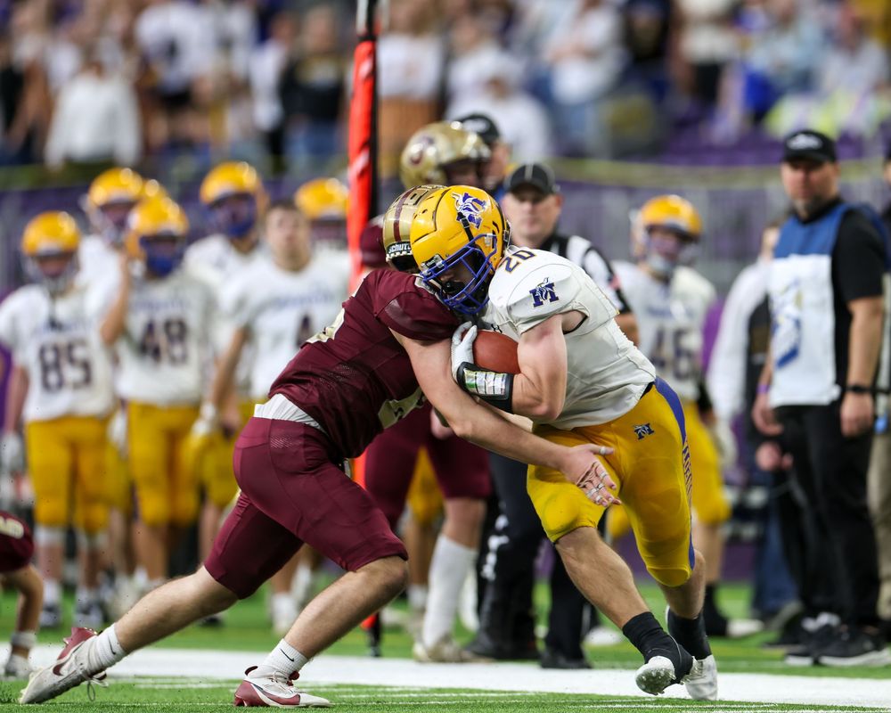 Two football players collide during an indoor game, one tackling the other who carries the ball in purple and gold uniforms.