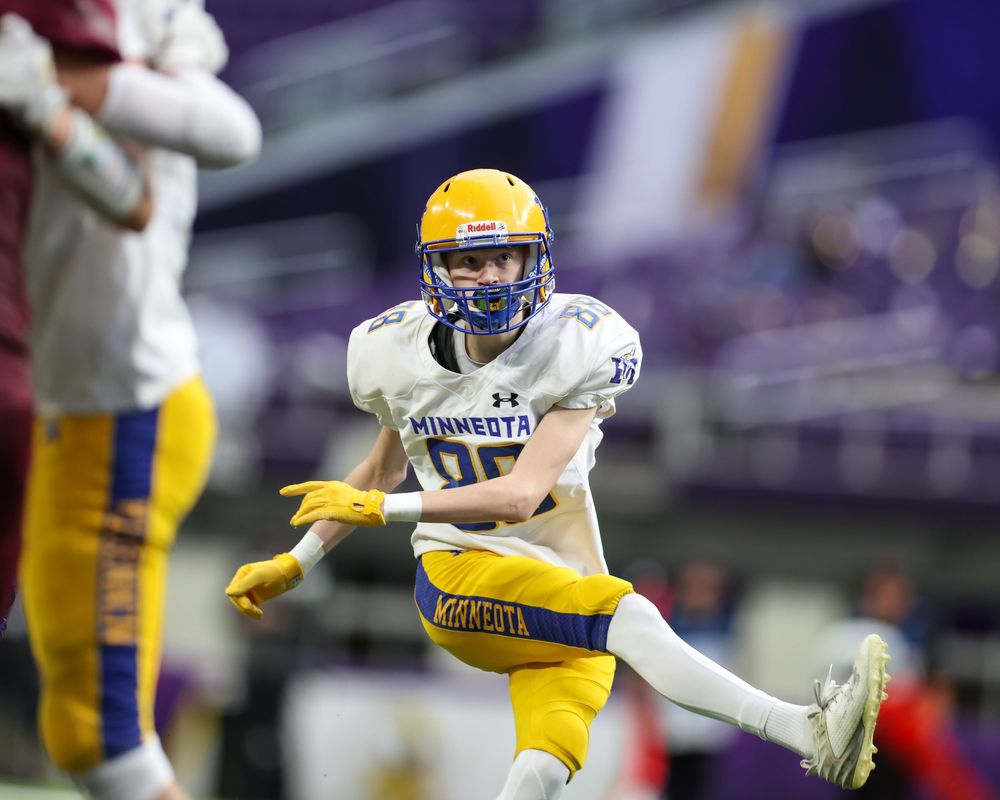 Minnesota football player in blue and gold uniform mid-play on indoor field during a high school championship game.