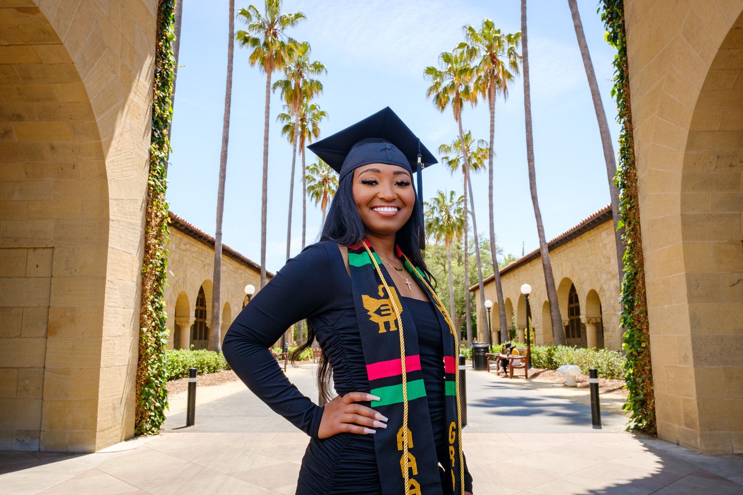 Smiling graduate in cap and gown with colorful stole poses between arched columns and palm trees on sunny campus.