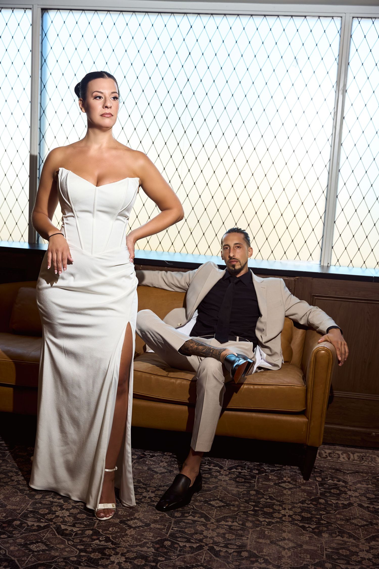 Elegant couple posing in upscale interior, woman in white strapless gown, man in beige suit on leather sofa.