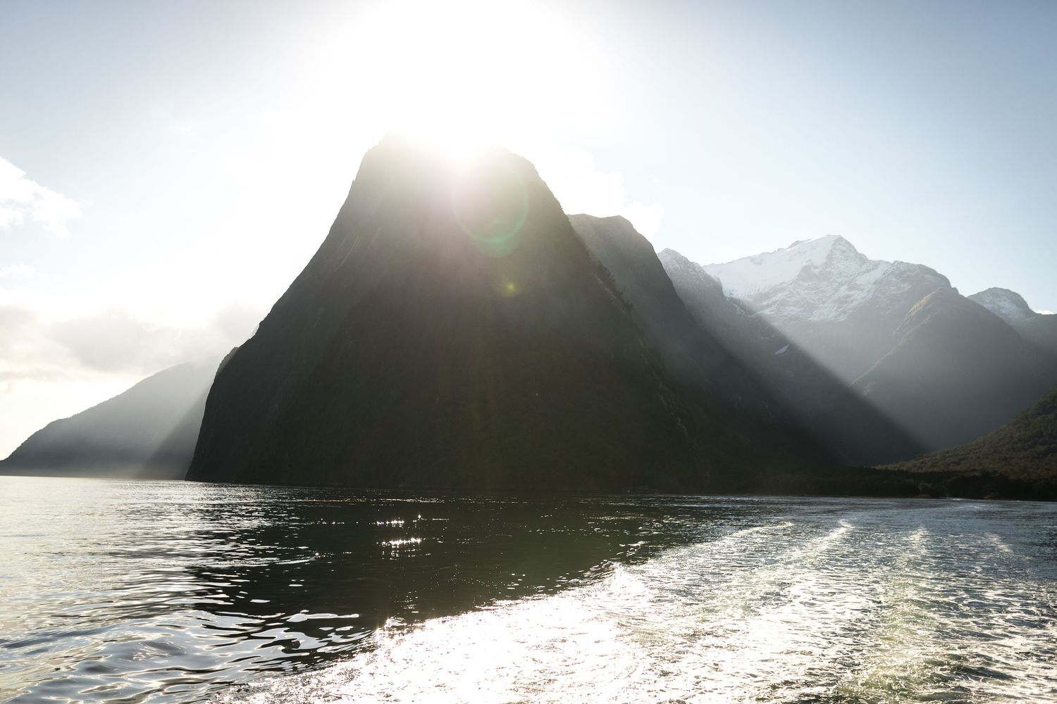 Sunlight bursts over a dramatic mountain peak reflected in calm fjord waters at Milford Sound, New Zealand.