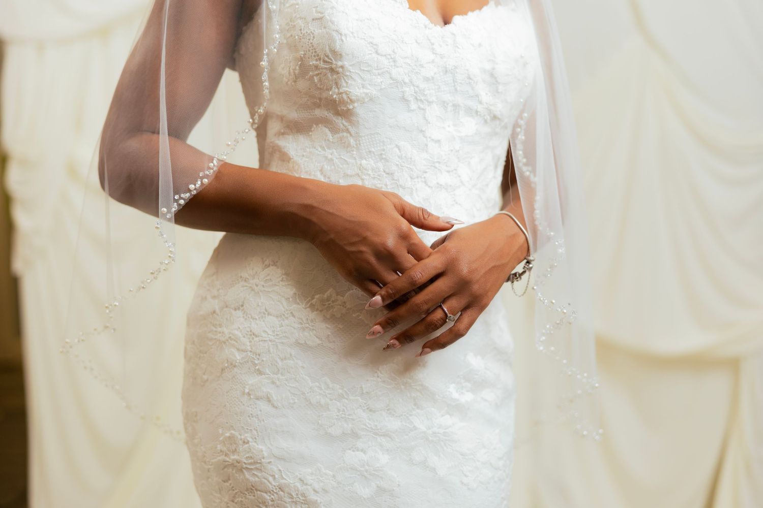 Bride in a strapless lace mermaid wedding dress with veil, hands clasped at waist, wearing bracelet and ring.