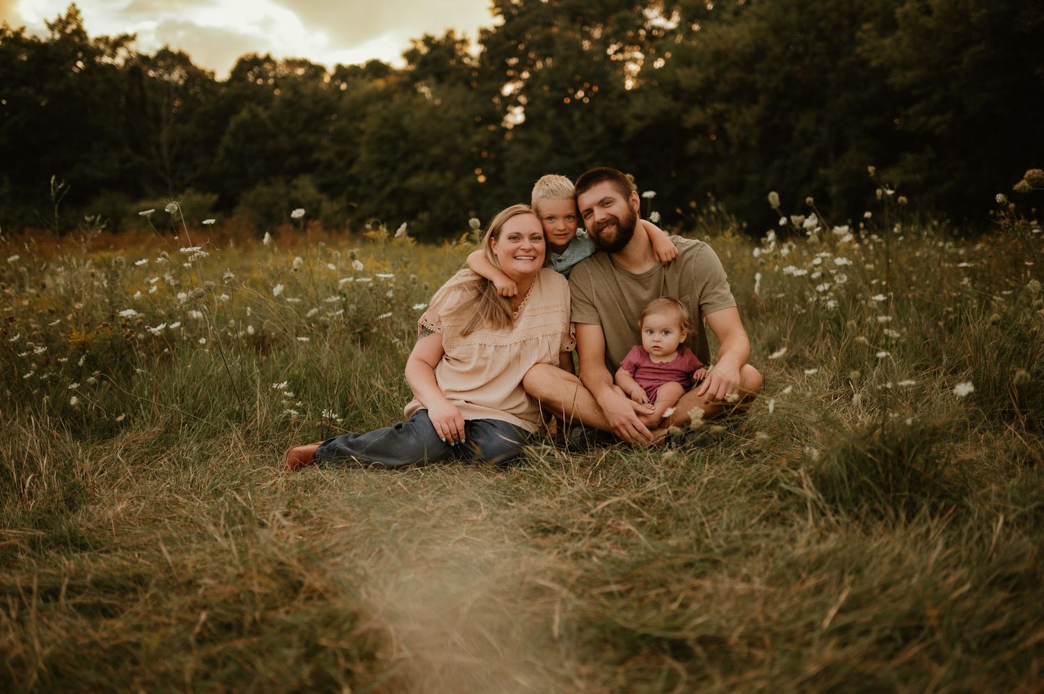 Family of four sitting in a sunlit wildflower meadow, smiling together at golden hour.
