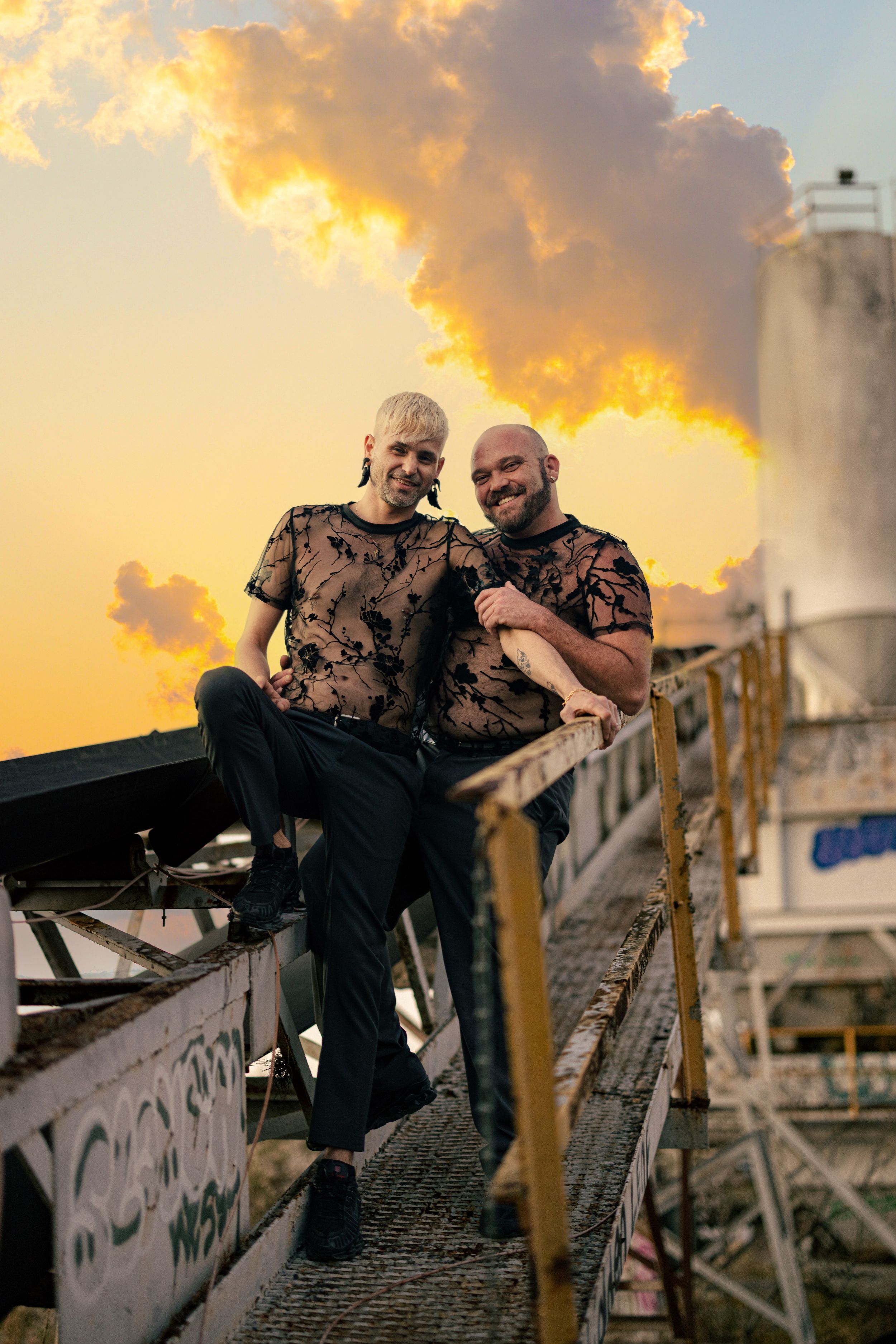 Two men in matching tie-dye shirts smile together on an industrial rooftop at golden hour sunset.