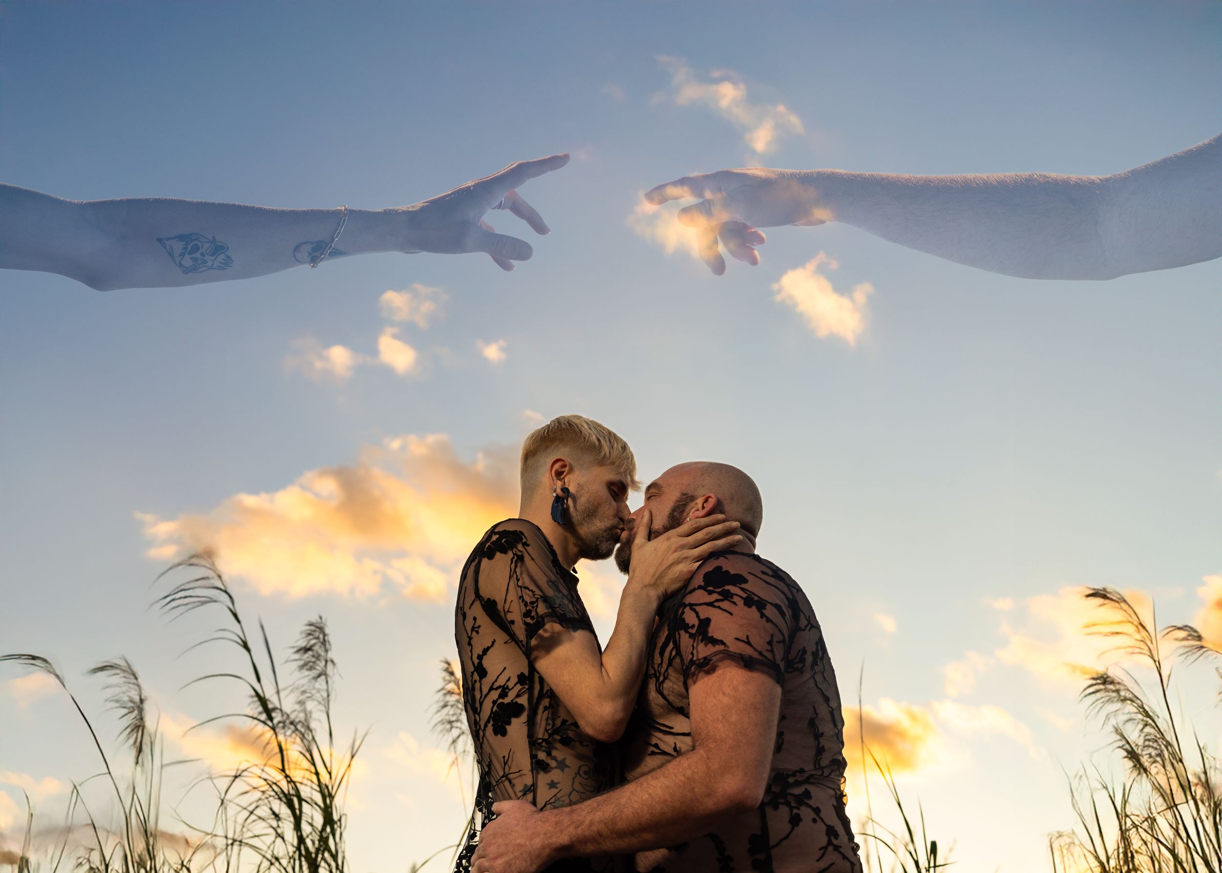 Two men share a romantic kiss outdoors at sunset with artistic hands reaching toward each other in the sky above.