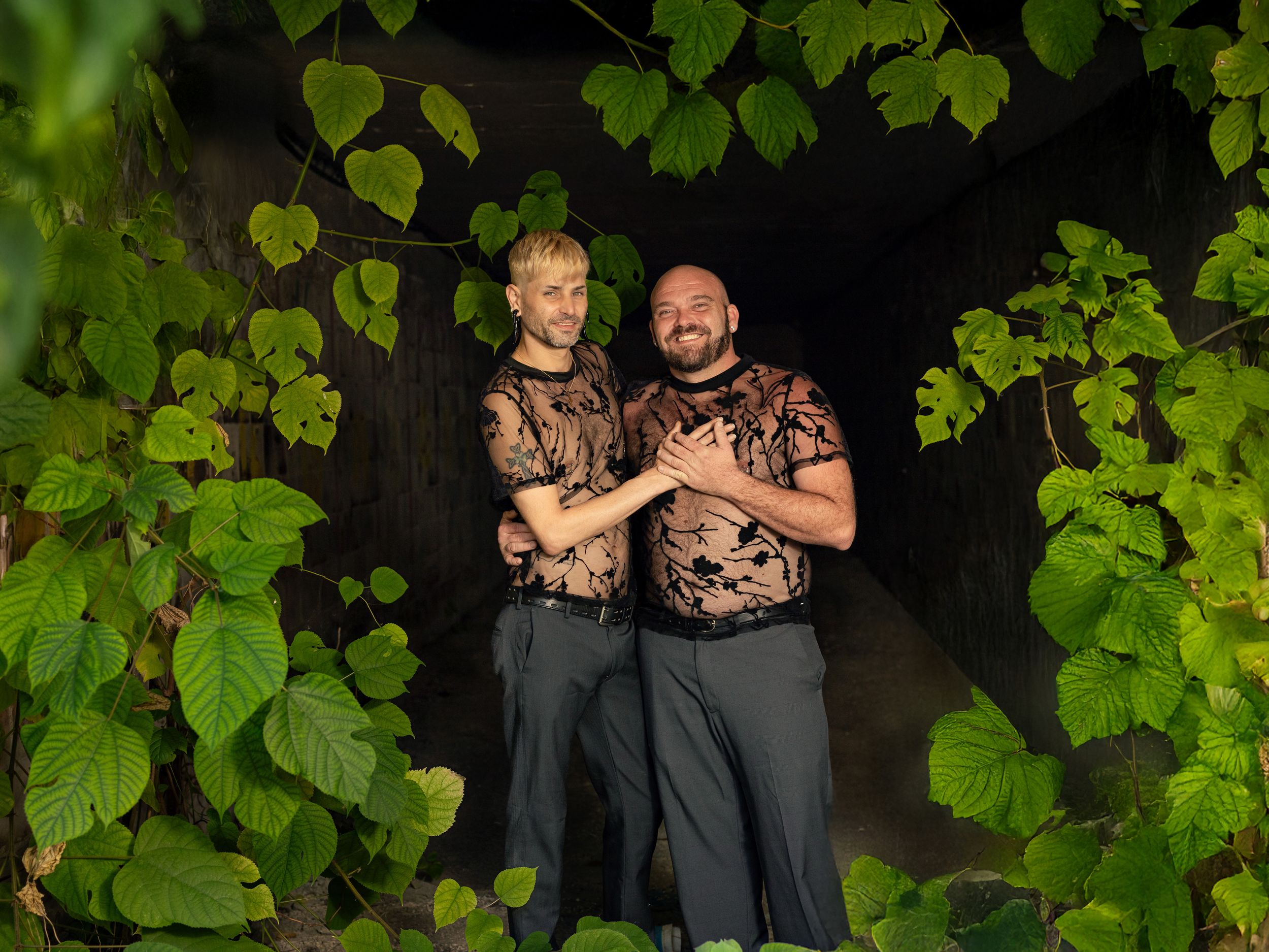 Two men in matching floral tops pose together in a lush green vine-covered archway.