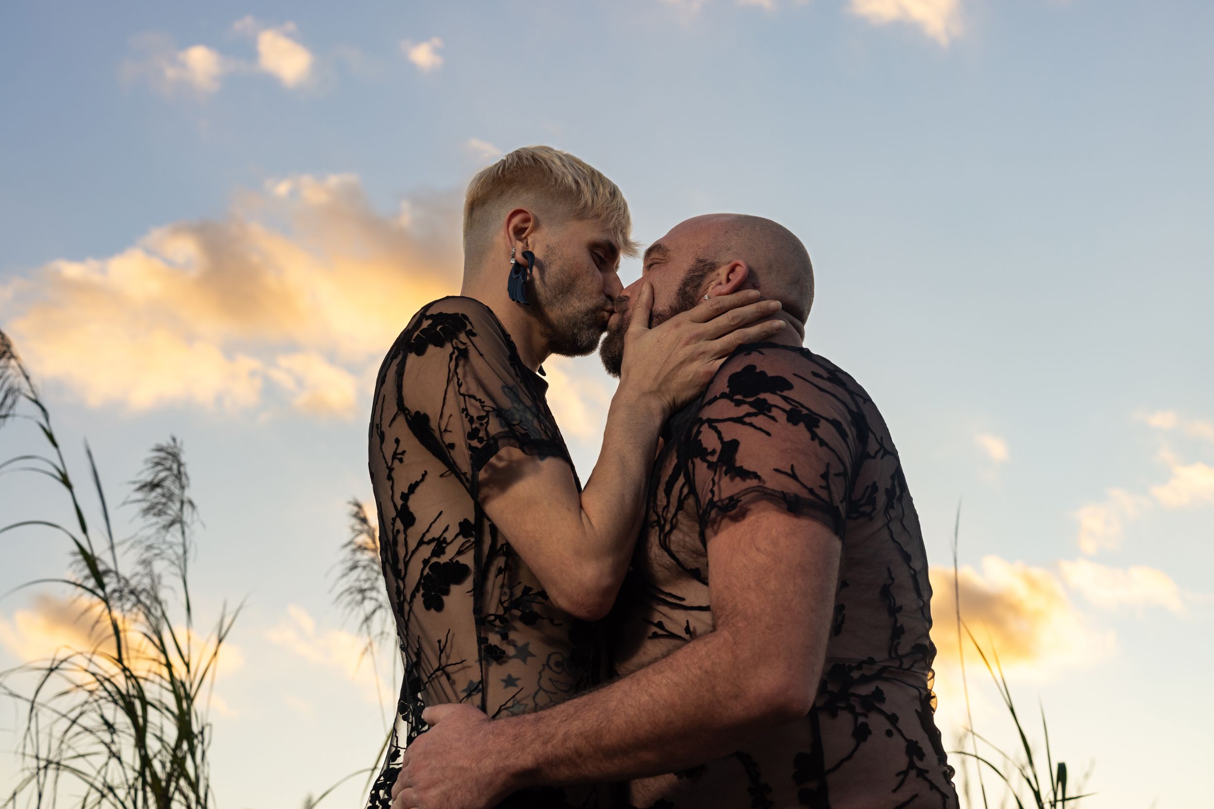 Two men sharing a romantic kiss outdoors at sunset, wearing matching black floral tops against a cloudy sky.