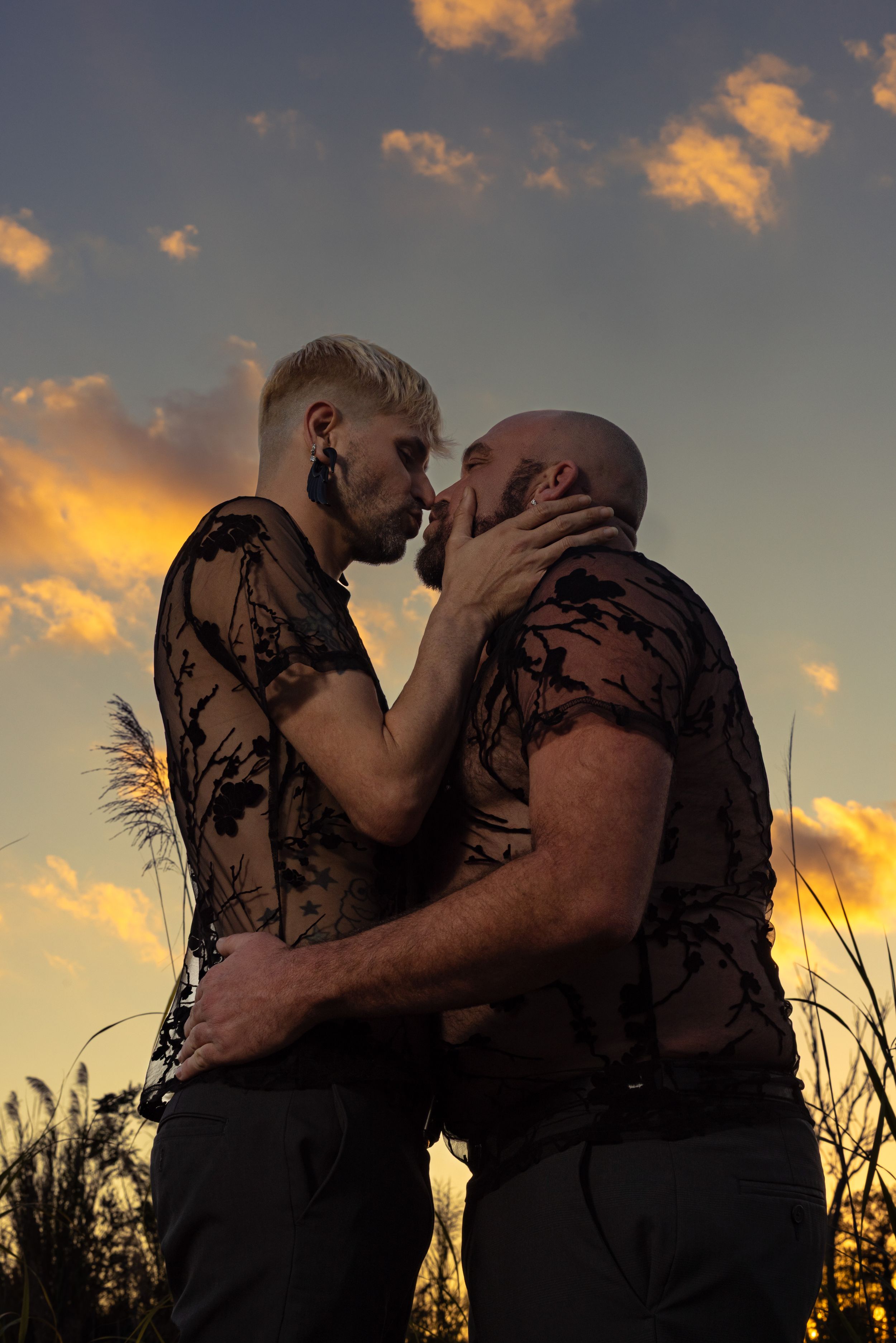 Two men sharing a romantic kiss at sunset with dramatic golden clouds in the background.