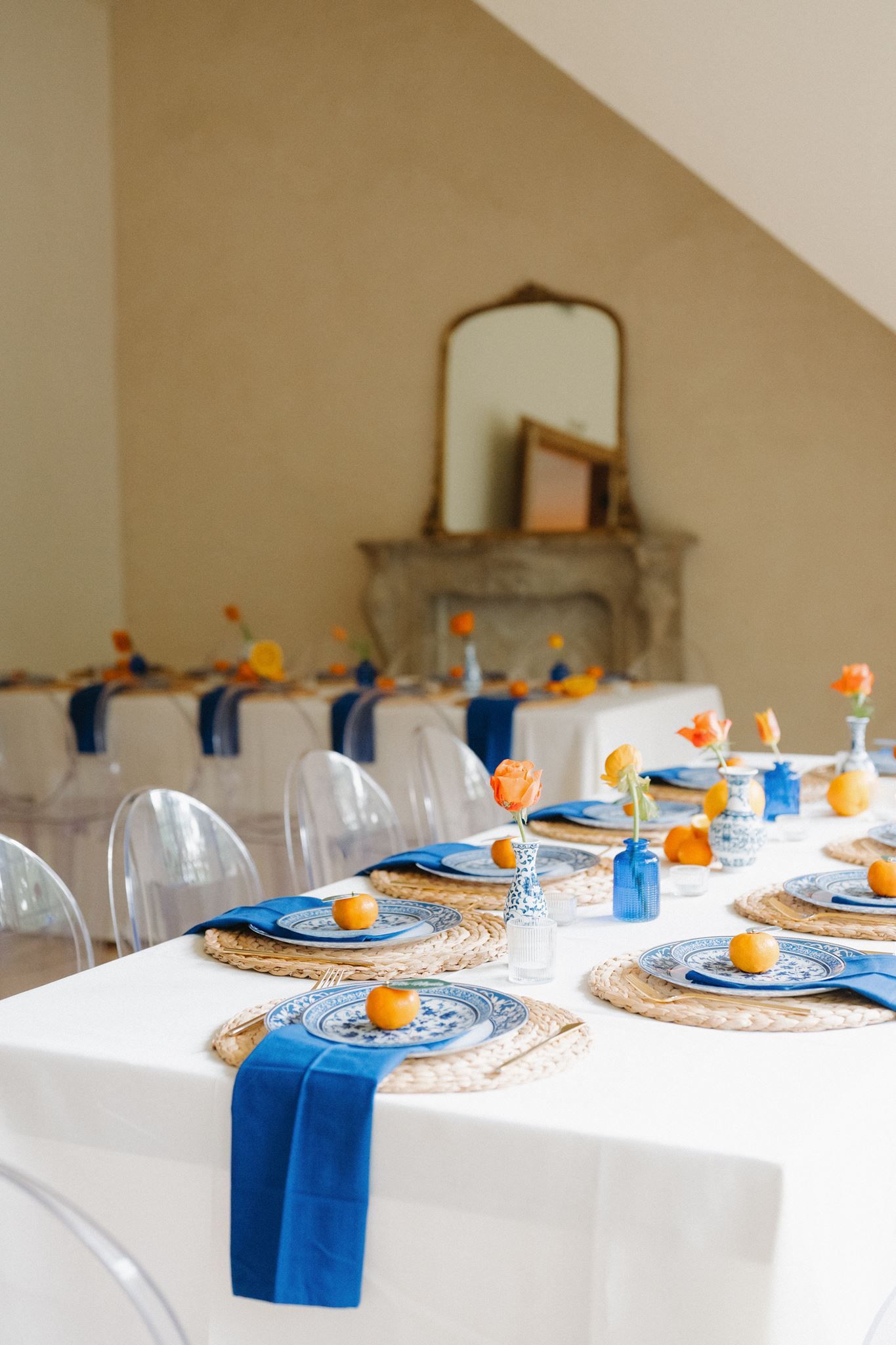 Elegant wedding reception table with blue napkins, wicker chargers, blue plates, orange flowers in blue vases, and ghost chairs.