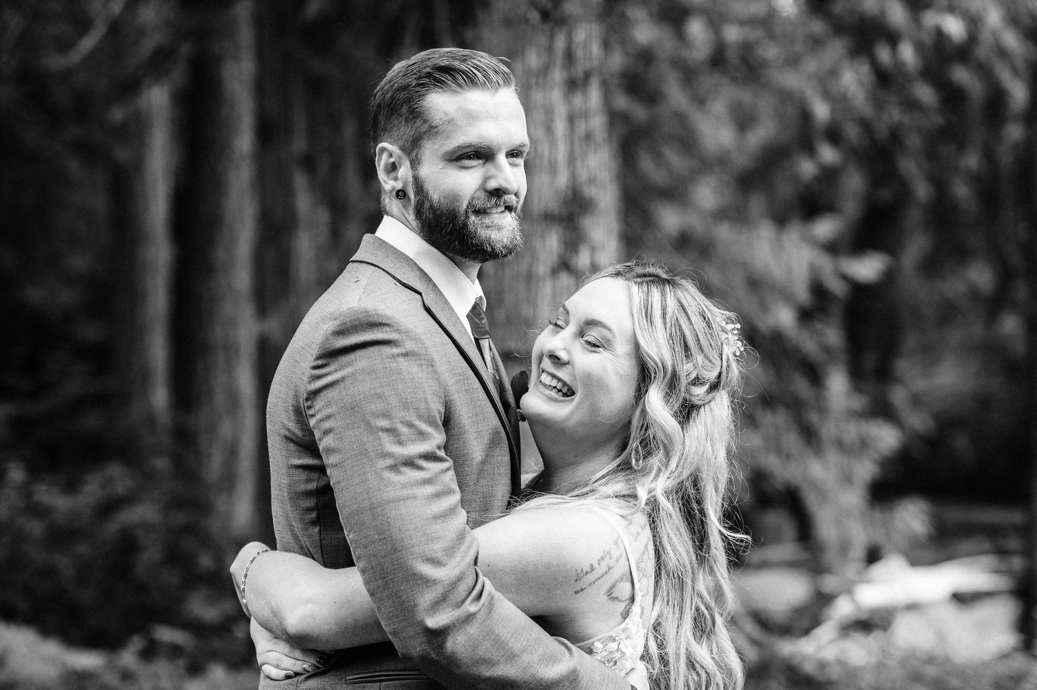 Black and white photo of a smiling couple embracing outdoors among tall trees, dressed in wedding attire.