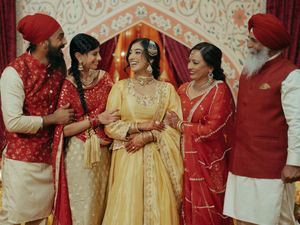 A Sikh bride in yellow lehenga smiles with family in red traditional attire at a vibrant Indian wedding ceremony.