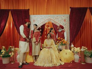 Indian wedding ceremony with bride in gold lehenga seated on ornate stage, groom and guests in red traditional attire.