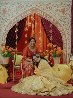 Bride in golden lehenga leans on mother in red saree at ornate Indian wedding mandap with marigold flowers.