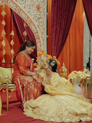 Bride in golden lehenga sits with elder woman in red suit during Indian wedding mehndi ceremony.