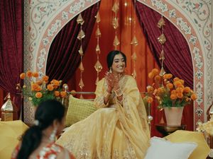 Bride in golden lehenga sits smiling during Indian wedding ceremony with marigold flowers and red drapes.