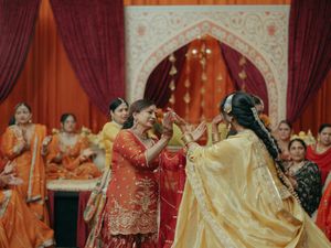 Two women in vibrant traditional Indian attire share a joyful moment at a colorful wedding ceremony.