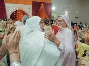 Elderly women in white dupattas embrace warmly at a vibrant Indian wedding celebration.