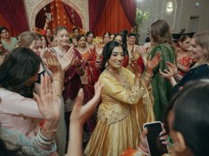 Bride in golden lehenga dances joyfully surrounded by guests clapping at Indian wedding mehndi.