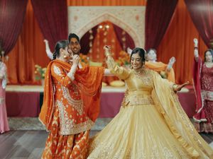 Bride in golden lehenga dances with woman in orange salwar at vibrant Indian wedding sangeet ceremony.