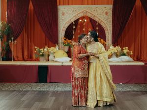 Two women in vibrant South Asian bridal attire embrace warmly at a decorated wedding venue with orange drapes.