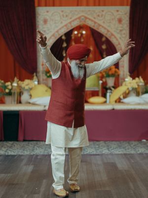 Elderly Sikh man in red turban and vest dances joyfully at Indian wedding celebration.