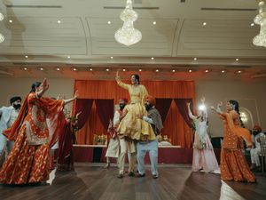 Guests perform a vibrant Bollywood dance at an Indian wedding reception with orange drapes and chandeliers.