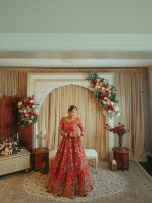 Indian bride in red lehenga stands before elegant floral arch backdrop at Sikh wedding ceremony.