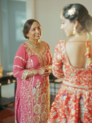 A smiling woman in a pink embroidered salwar kameez with gold jewelry speaks with a bride in a red lehenga.