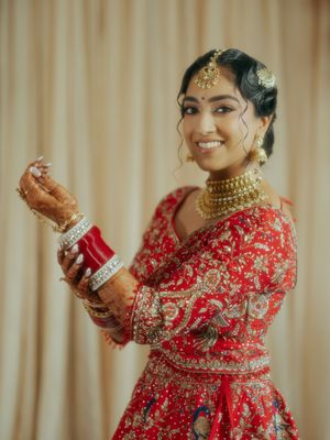 Indian bride in red embroidered lehenga with gold jewelry and mehndi, smiling elegantly.