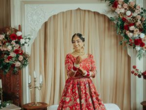 Smiling Indian bride in red embroidered lehenga poses before decorative arch with floral arrangements.