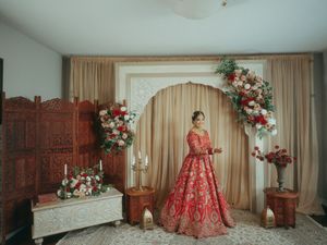 Bride in red embroidered lehenga standing before floral arch with roses and candles in elegant indoor setting.