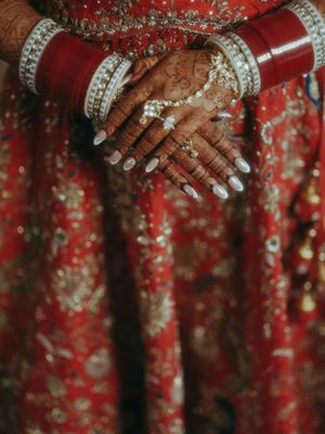 Close-up of Indian bride's hands adorned with henna, white nail polish, bangles, and rings on red embroidered lehenga.