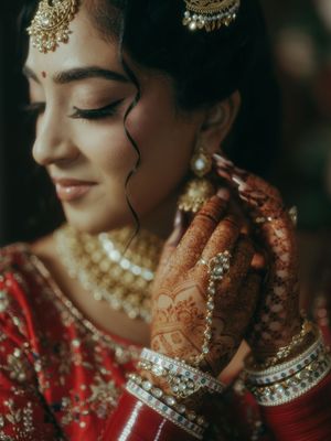 Indian bride in red lehenga adjusting gold earring with mehndi-adorned hands and traditional bridal jewelry.
