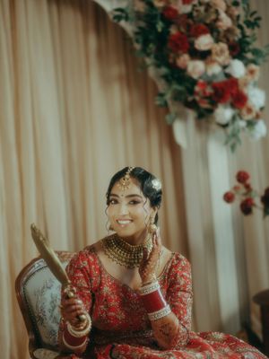 A smiling Indian bride in red lehenga with gold jewelry and maang tikka holds a decorative item while seated near floral decor.