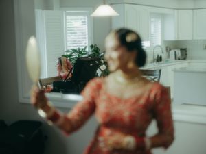 Woman in red lace dress holding a hand mirror in a bright kitchen interior.