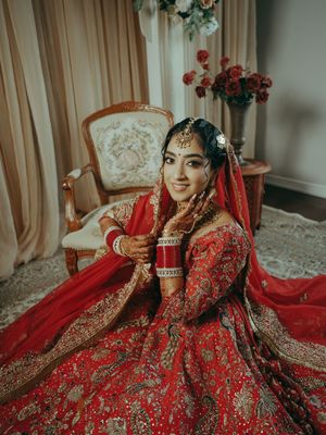 Bride in red and gold embroidered lehenga with dupatta and bridal jewelry seated elegantly indoors.